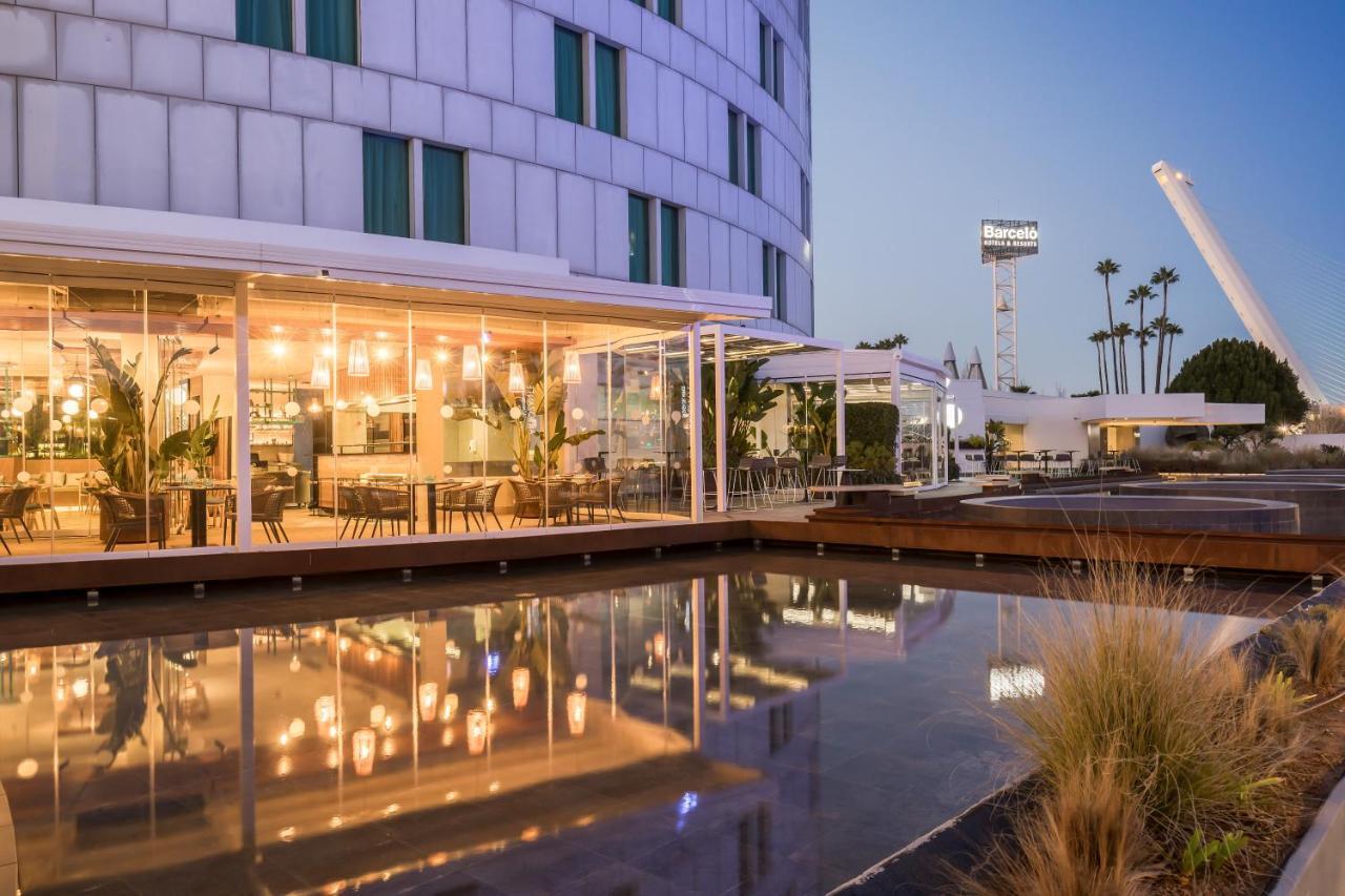 Barcelo Sevilla Renacimiento Esterno foto The photo shows a modern architectural building, likely a hotel or a restaurant, with large windows and a contemporary design. There’s an outdoor area with a dining space, featuring stylish lighting and greenery. In the foreground, there’s a reflecti