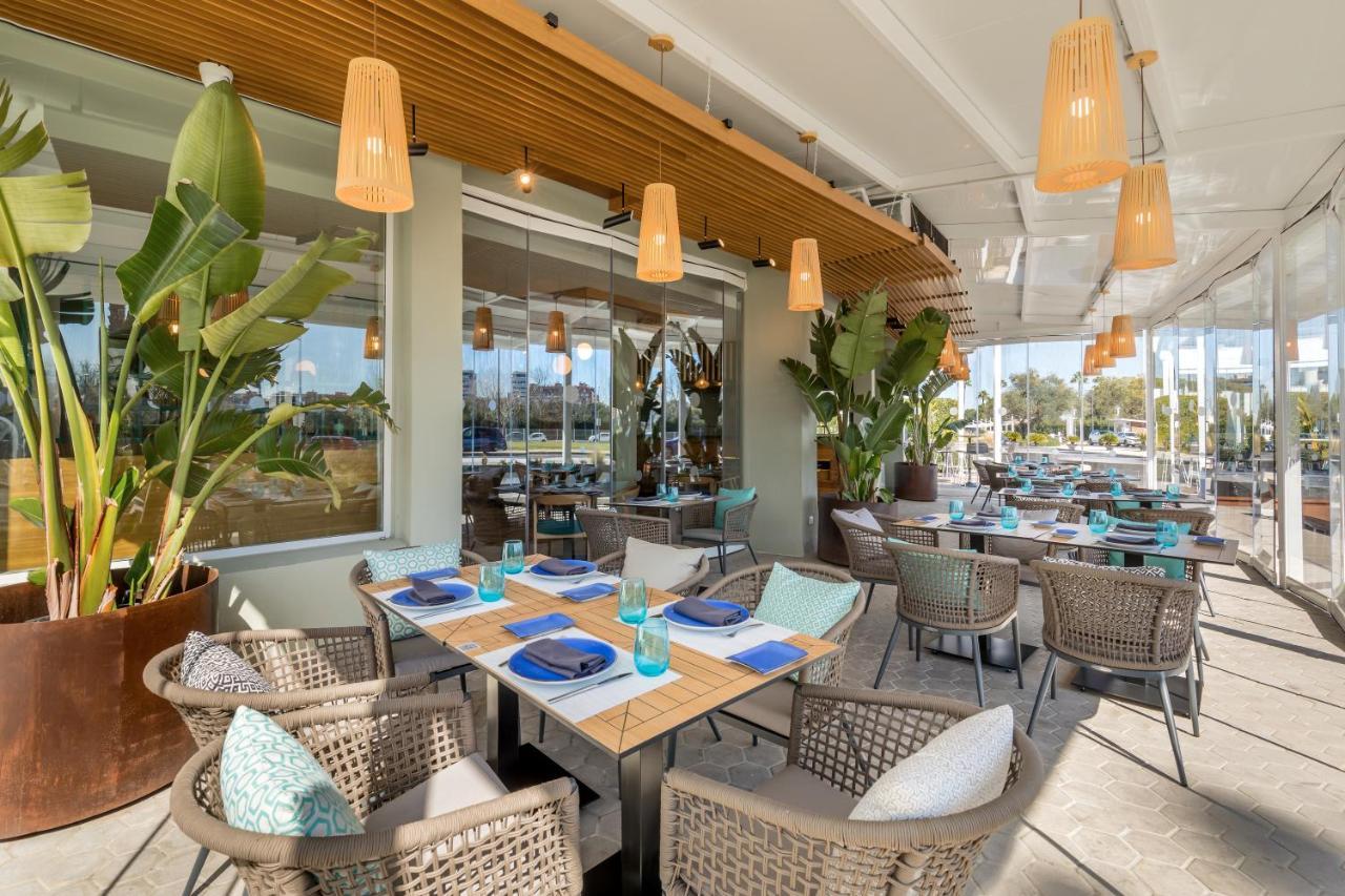 Barcelo Sevilla Renacimiento Esterno foto The photo shows a well-decorated restaurant interior. There are several dining tables arranged with blue napkins and turquoise glasses. The seating consists of woven chairs, providing a cozy atmosphere. Large windows allow natural light to fill the s