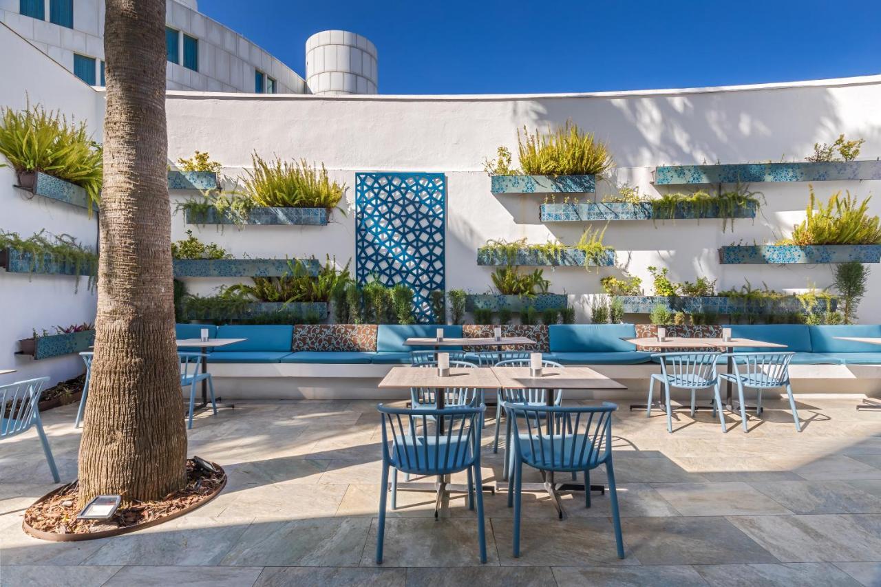 Barcelo Sevilla Renacimiento Esterno foto The photo shows a charming outdoor café or restaurant area. It features several tables with blue chairs arranged in an inviting setting. Behind the tables, there’s a wall adorned with vertical planters showcasing various green plants, adding a touch 
