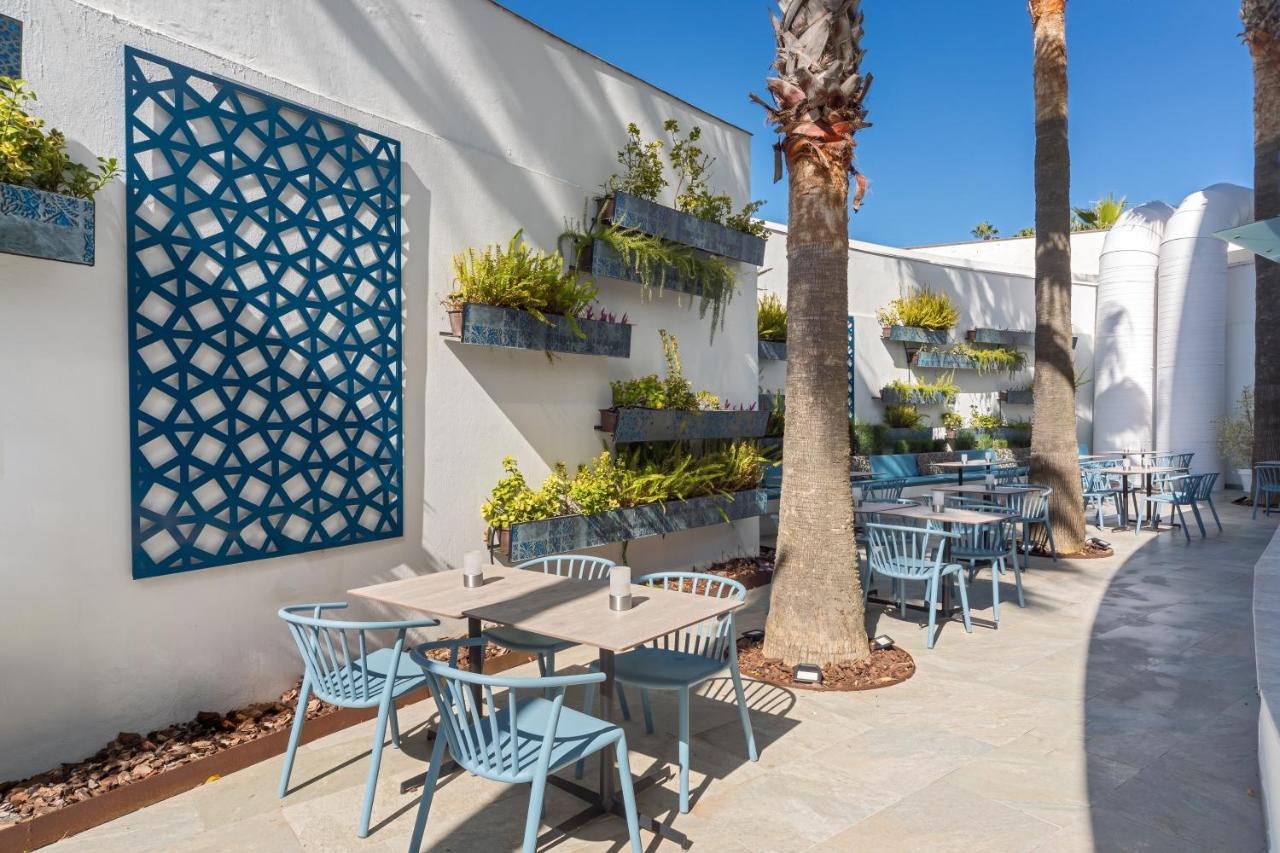 Barcelo Sevilla Renacimiento Esterno foto The photo depicts a modern outdoor dining area featuring light blue chairs and tables arranged neatly. The walls are painted white, and there is a decorative blue lattice panel on one side. Vertical planters with greenery are mounted on the walls, ad