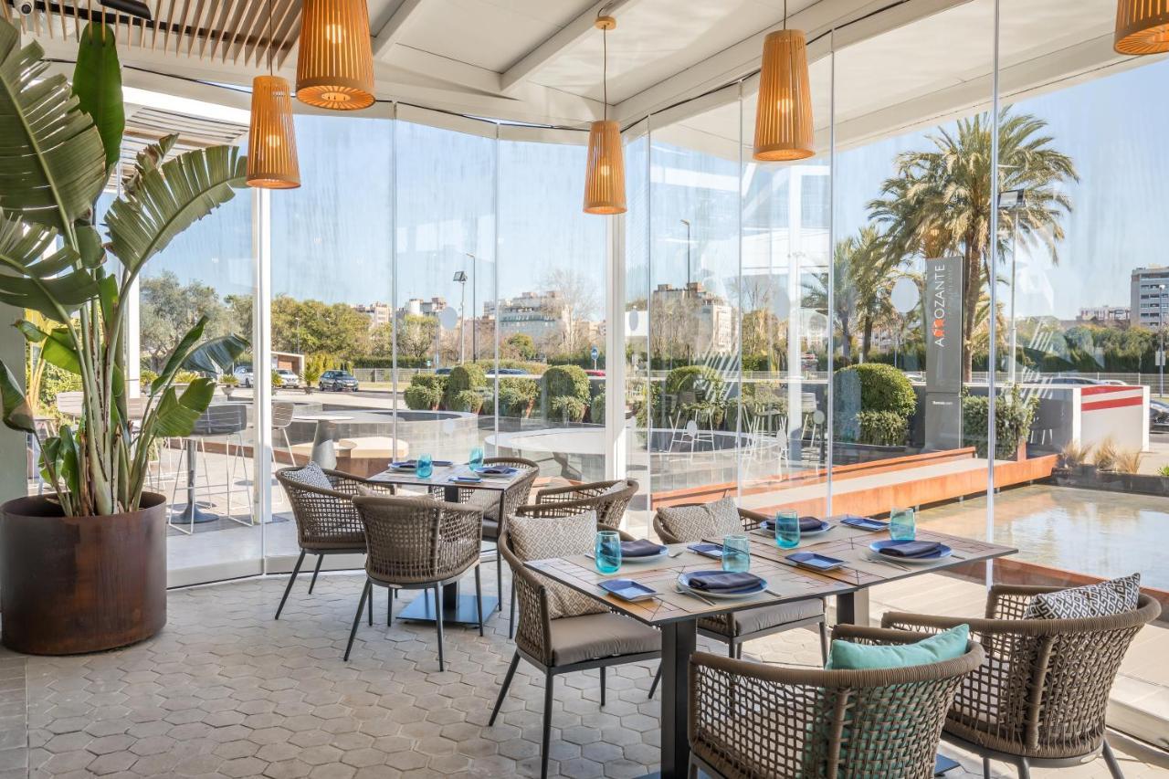 Barcelo Sevilla Renacimiento Esterno foto The photo depicts a modern restaurant interior featuring a bright and airy ambiance. There are several tables set up with wicker chairs, and each table is adorned with blue glassware. The space has large glass windows that provide a view of lush gree