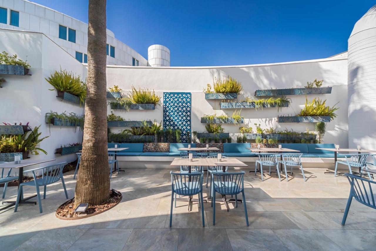 Barcelo Sevilla Renacimiento Esterno foto The photo shows a serene outdoor space, likely a café or restaurant. The area features multiple tables and chairs arranged on a tiled floor, complemented by walls adorned with greenery. There are shelves mounted on the walls filled with various plant