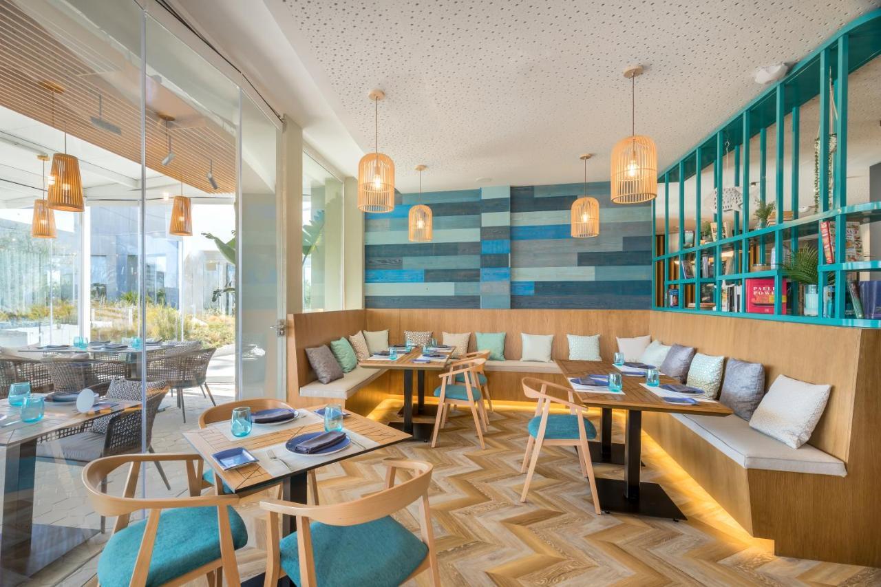 Barcelo Sevilla Renacimiento Esterno foto The image shows a modern and stylish restaurant interior. It features a combination of comfortable seating, including a wooden L-shaped bench adorned with colorful cushions. The decor displays a mix of pastel colors on the walls, particularly in shad