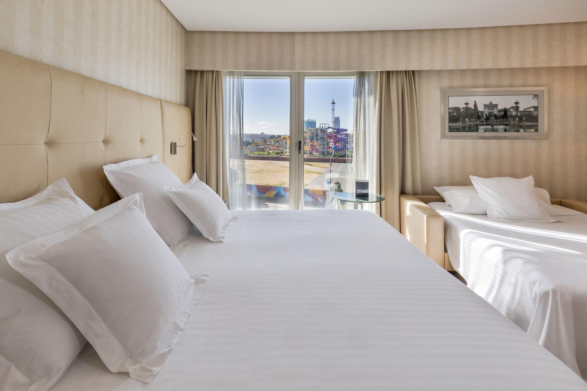 Barcelo Sevilla Renacimiento Esterno foto The photo shows a modern hotel room featuring a large bed with white bedding and several fluffy pillows. The walls have a subtle pattern, and there are large windows covered with light curtains that let in natural light. Through the windows, a view o