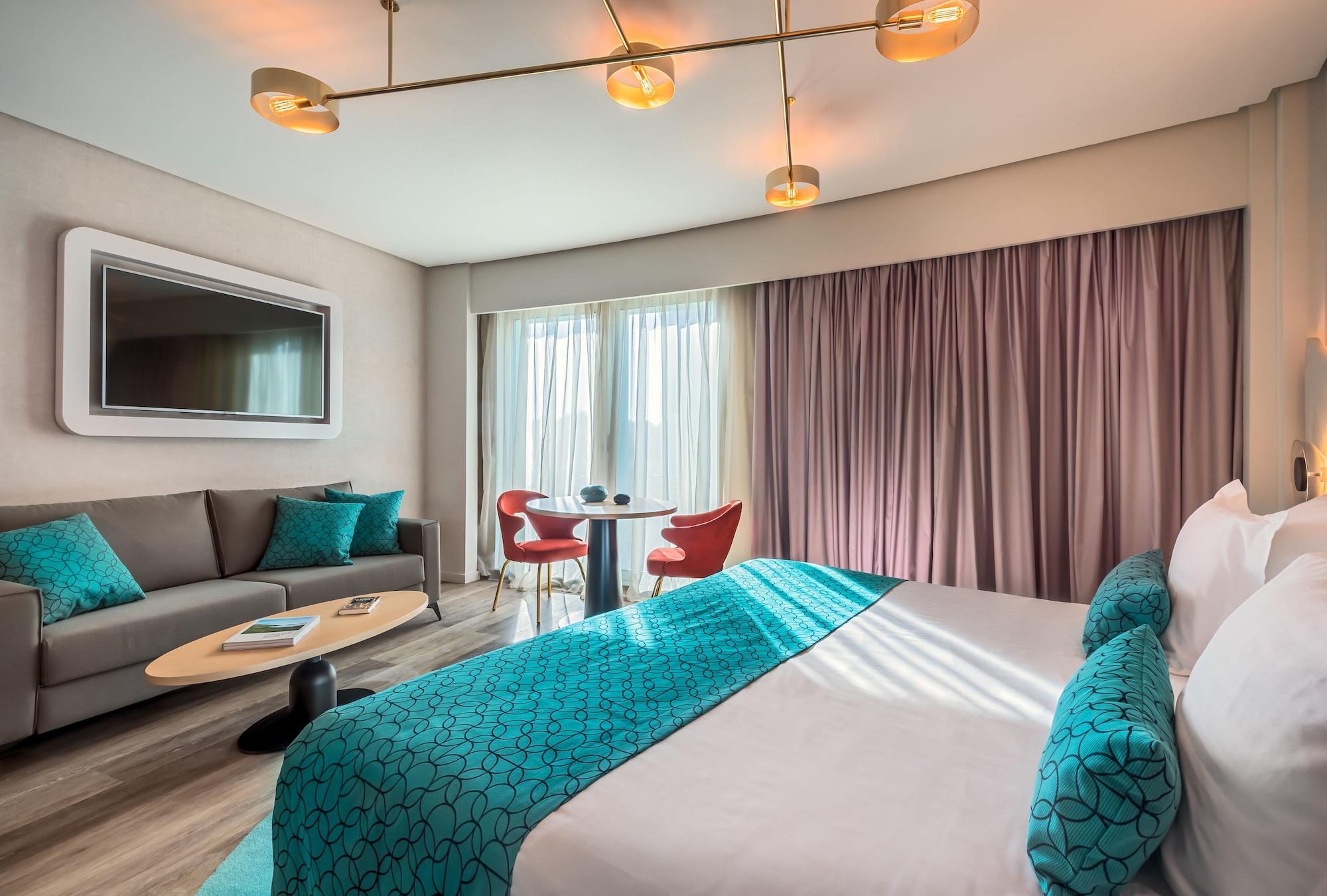Barcelo Sevilla Renacimiento Esterno foto The photo shows a modern hotel room featuring a large bed with a turquoise decorative throw and pillows. There’s a stylish sofa in a neutral tone, complemented by turquoise cushions. A small round table with red chairs sits nearby, creating a cozy di