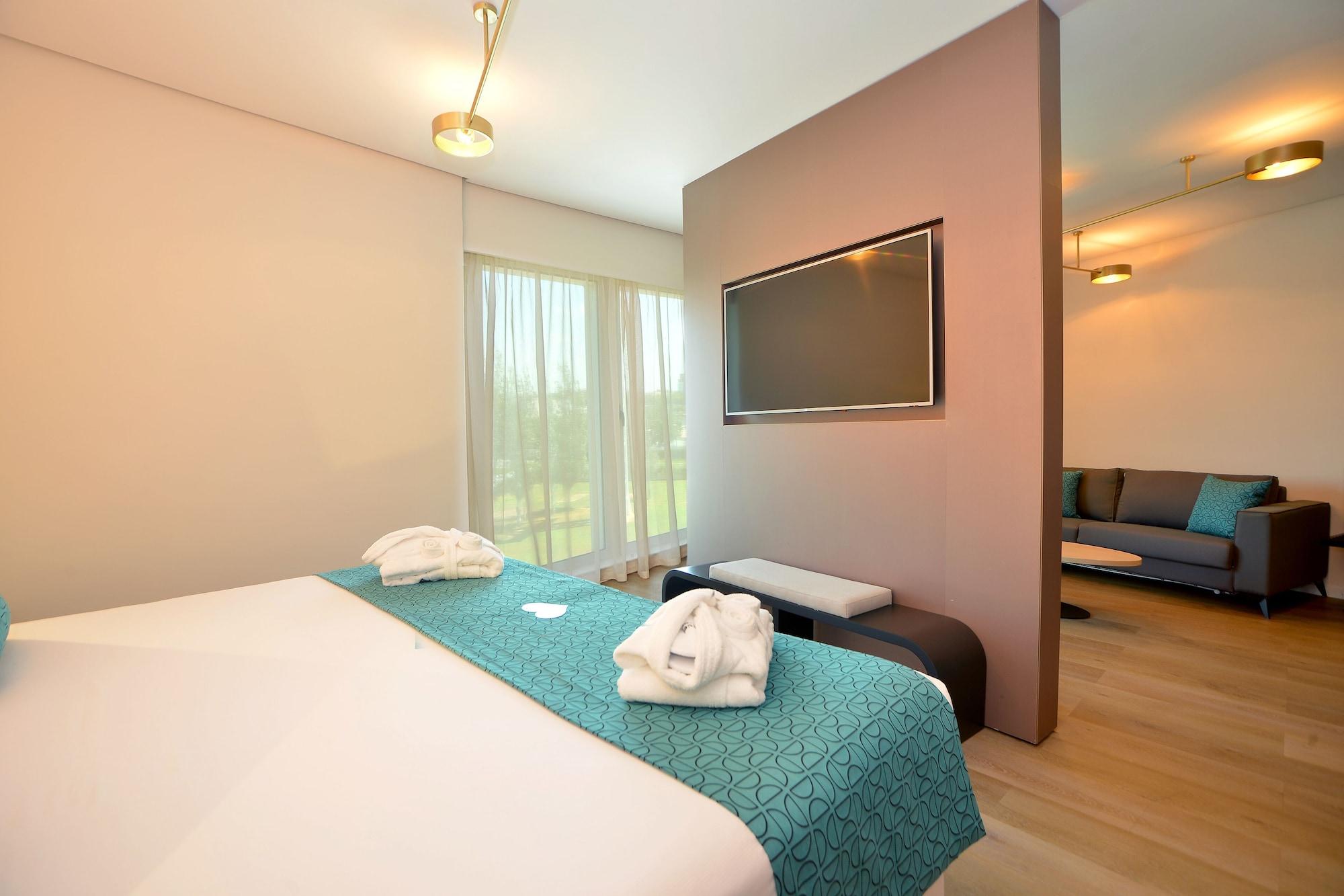 Barcelo Sevilla Renacimiento Esterno foto The photo shows a modern hotel room featuring a double bed with a light blue decorative blanket. There are neatly arranged white towels on the bed. A wall-mounted flat-screen TV is visible, along with a small seating area that includes a sofa. The ro