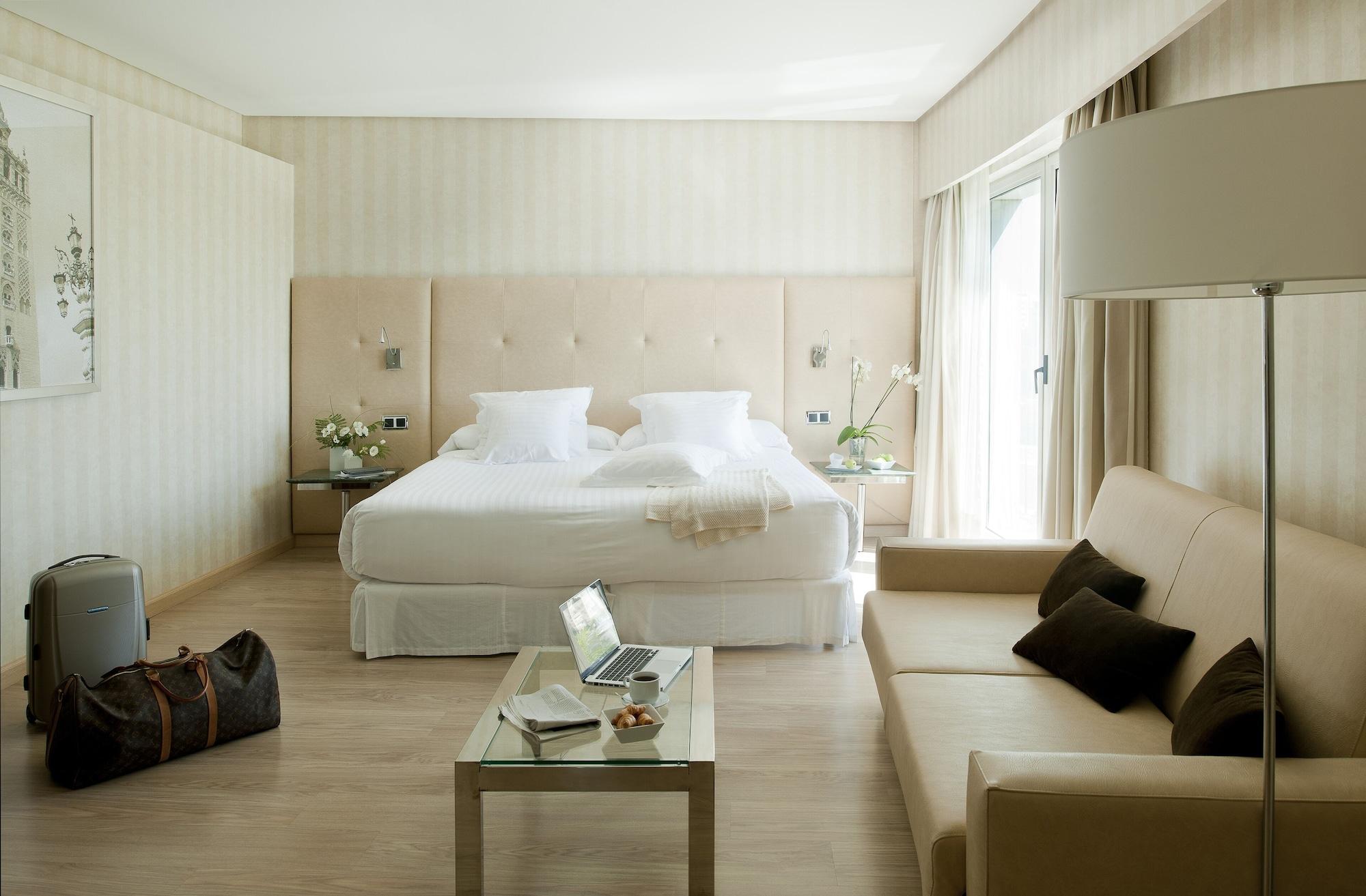Barcelo Sevilla Renacimiento Esterno foto The photo shows a modern and elegantly decorated hotel room. It features a large bed with a soft headboard and crisp white bedding. Beside the bed, there are small tables with decorative items, including flowers. A comfortable beige sofa is placed in