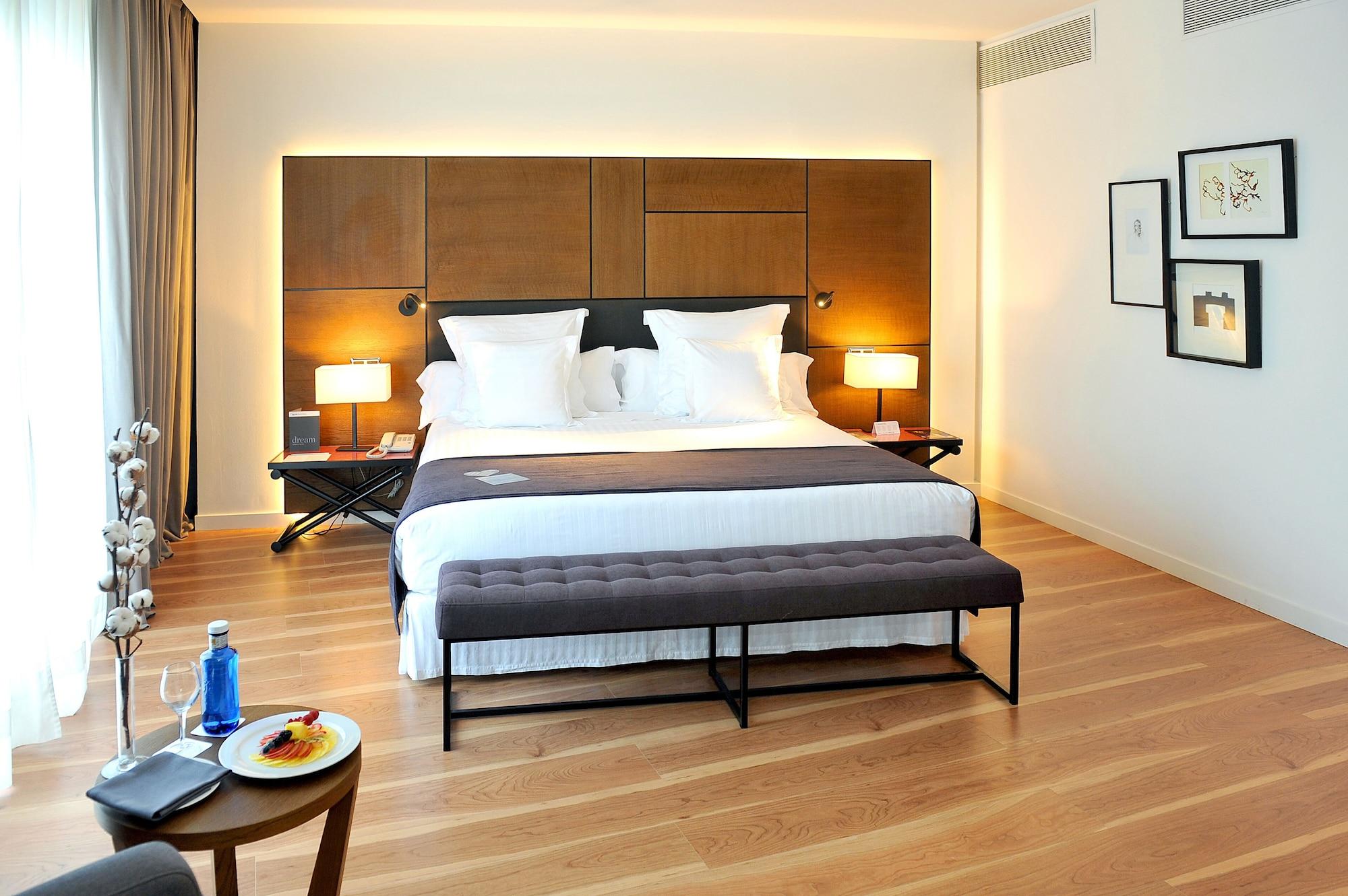 Barcelo Sevilla Renacimiento Esterno foto The photo shows a modern hotel bedroom. It features a large bed with white pillows and a neatly made blanket. On either side of the bed are lamps, providing soft lighting. A bench with a dark upholstery sits at the foot of the bed. The room has woode