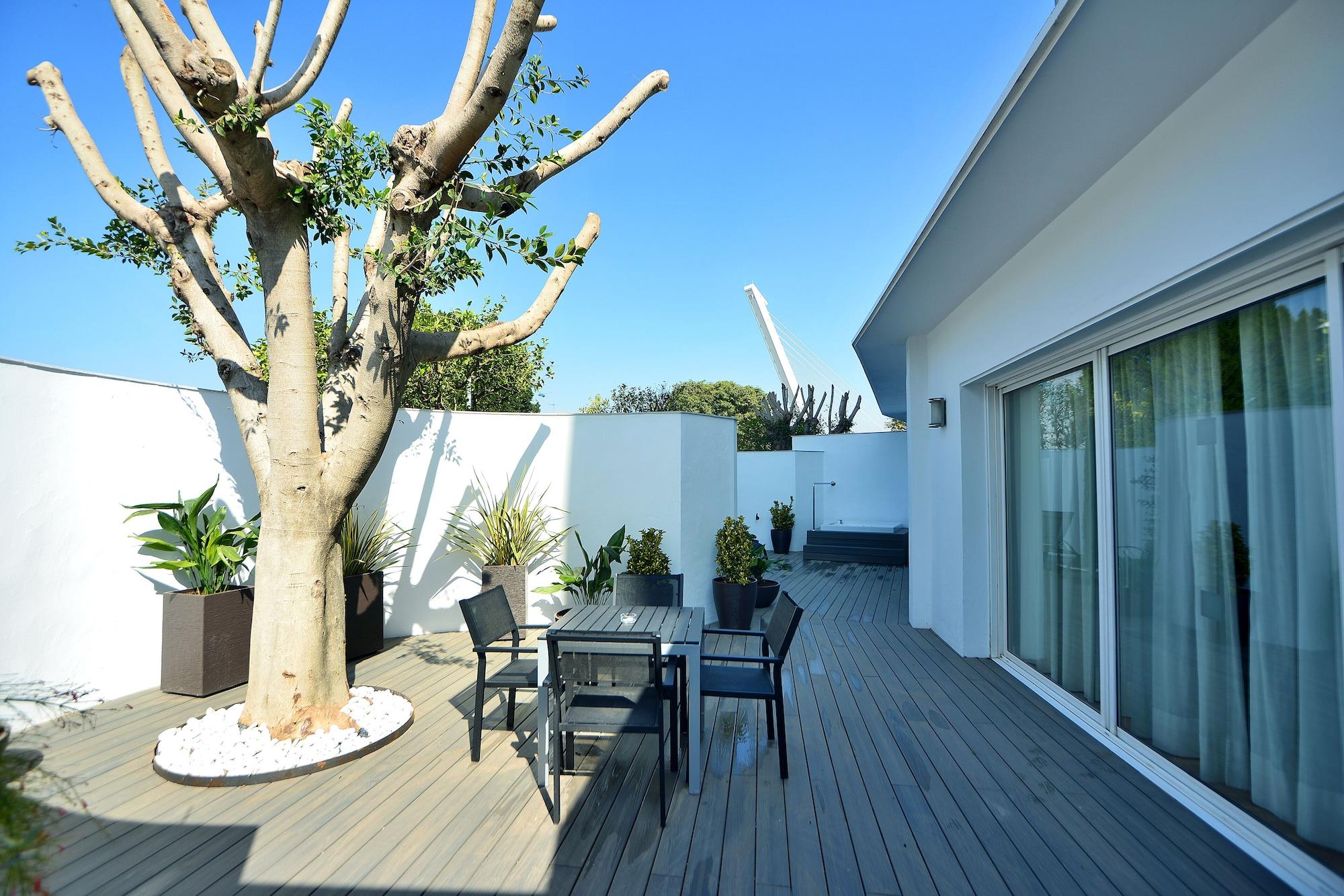 Barcelo Sevilla Renacimiento Esterno foto The photo shows a modern outdoor area featuring a wooden deck. There is a large tree in a circular bed of white stones, surrounded by potted plants. A table and four chairs are set up, creating a seating area. The backdrop includes a white wall and a