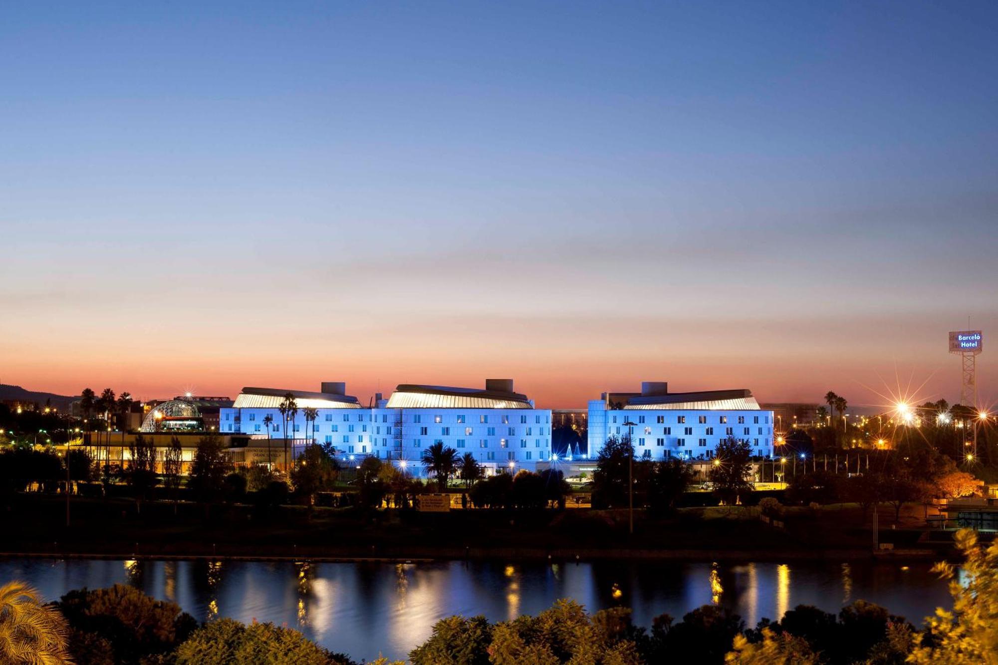 Barcelo Sevilla Renacimiento Esterno foto The photo shows modern buildings illuminated in blue against a twilight sky. The buildings are situated near a body of water, which reflects the blue lights. The scene suggests a serene and stylish atmosphere, with a mix of urban designs and natural 