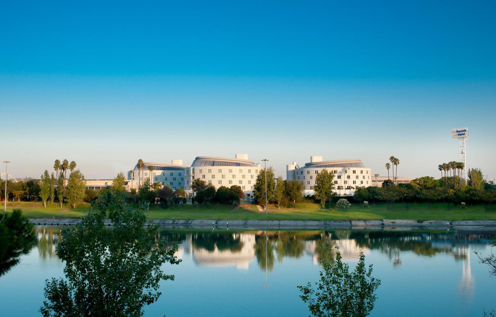 Barcelo Sevilla Renacimiento Esterno foto The photo shows a calm lakeside scene featuring several buildings in a modern architectural style. These buildings are situated along the water's edge, reflecting in the lake's surface. Surrounding the buildings, there are lush trees and greenery, co