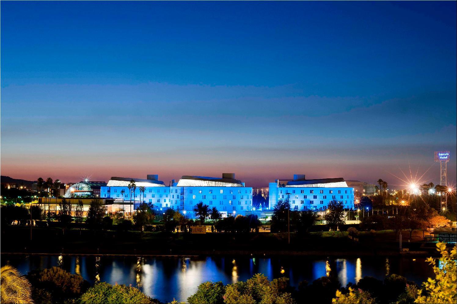 Barcelo Sevilla Renacimiento Esterno foto The photo depicts a modern building complex illuminated in blue lights, set against a twilight sky. The buildings are mirrored in the calm water of a nearby body of water, creating a serene and picturesque scene. The background features a gradient of