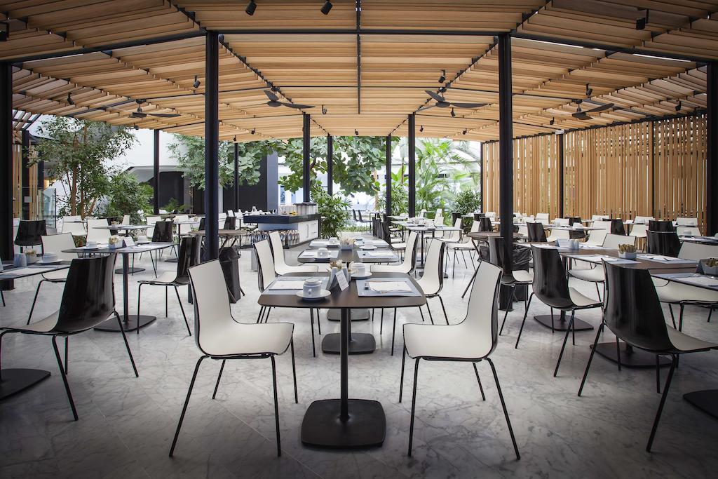 Barcelo Sevilla Renacimiento Esterno foto The image shows a modern restaurant interior with a spacious layout. There are numerous tables set up with white chairs and black tabletops arranged neatly in a well-lit space. The restaurant features a ceiling with wooden slats that allows natural l