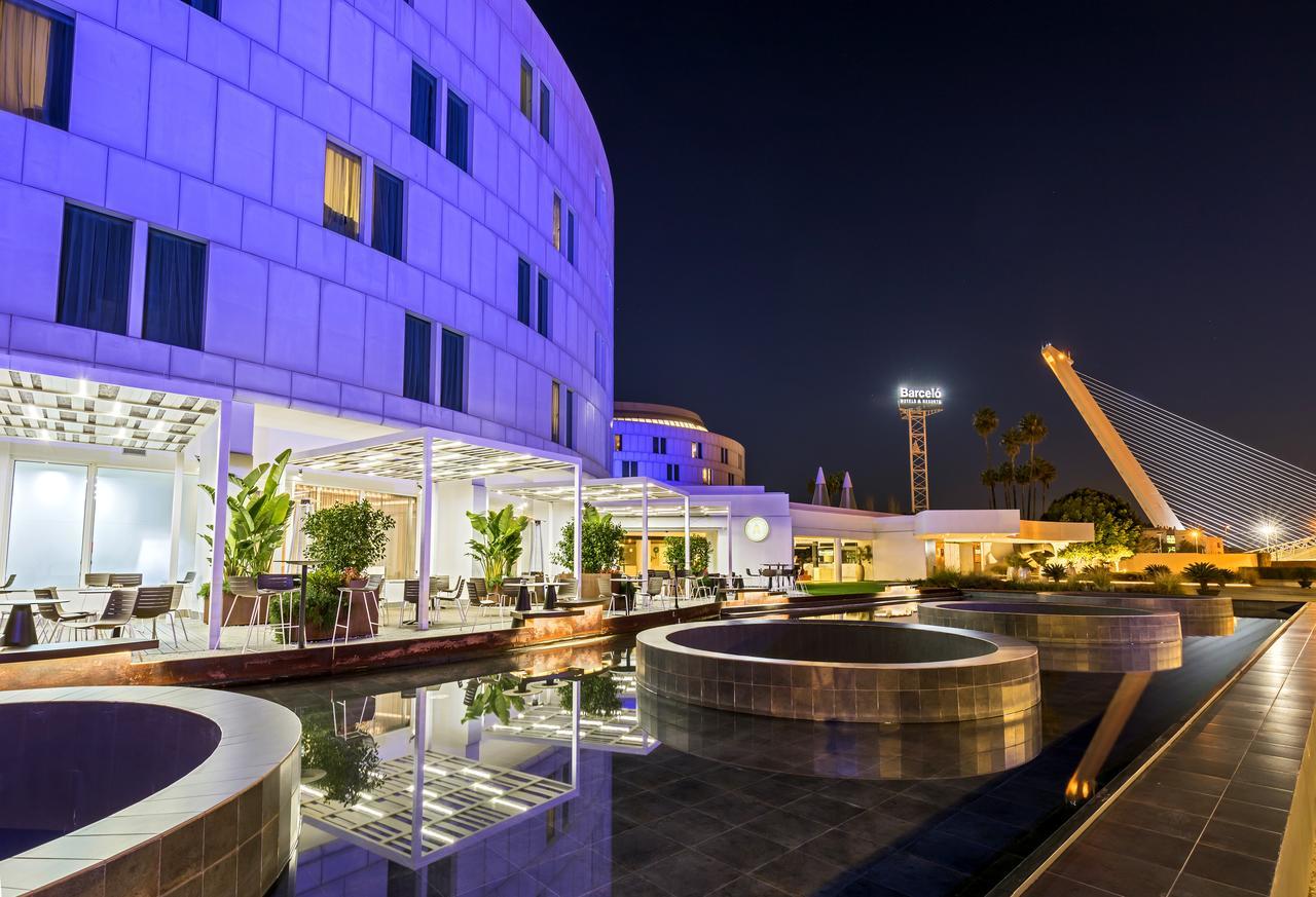 Barcelo Sevilla Renacimiento Esterno foto The photo features a modern architectural scene at night, showcasing a sleek building illuminated with purple lighting. In the foreground, there are round water features or pools reflecting the lights and the structure. There are also green plants ar