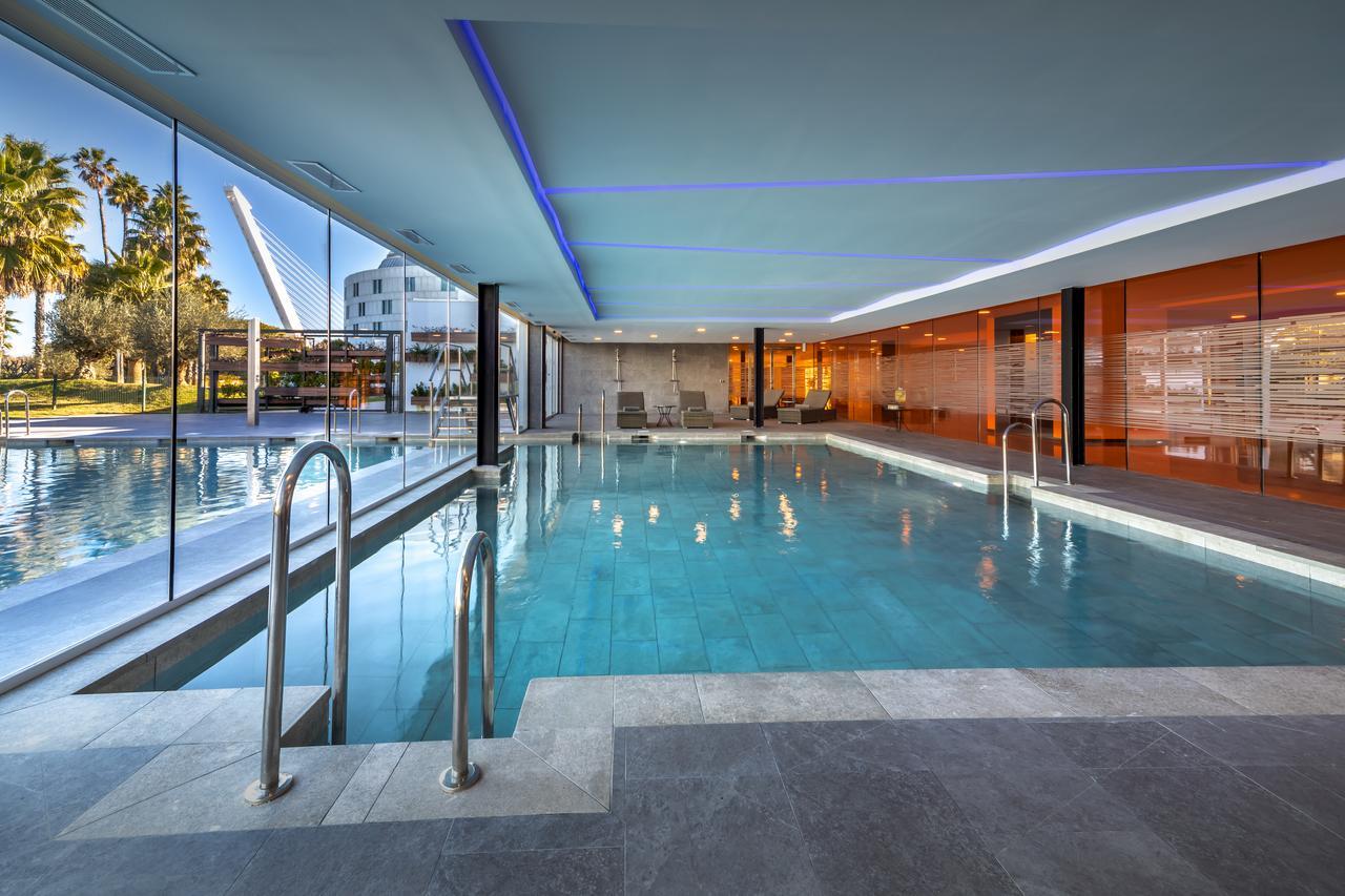 Barcelo Sevilla Renacimiento Esterno foto The photo shows a modern indoor swimming pool area with large glass walls allowing natural light to fill the space. The pool appears to have a clear, inviting blue water, and there are metallic ladders at one end for easy access. The surrounding area
