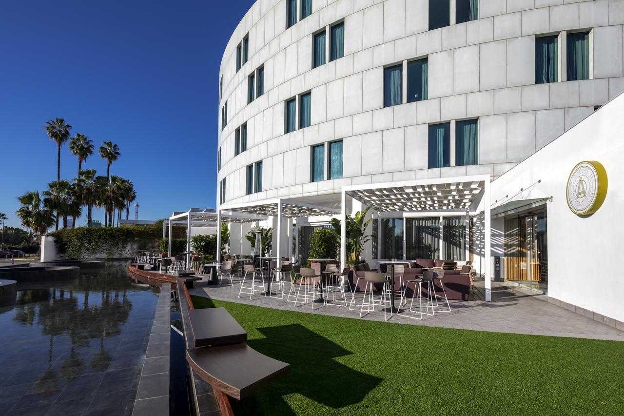 Barcelo Sevilla Renacimiento Esterno foto The photo shows a modern, circular building with a sleek, white facade. In the foreground, there's a patio area with outdoor seating, featuring stylish tables and chairs under a pergola. Surrounding the area are well-manicured green lawns and palm tr
