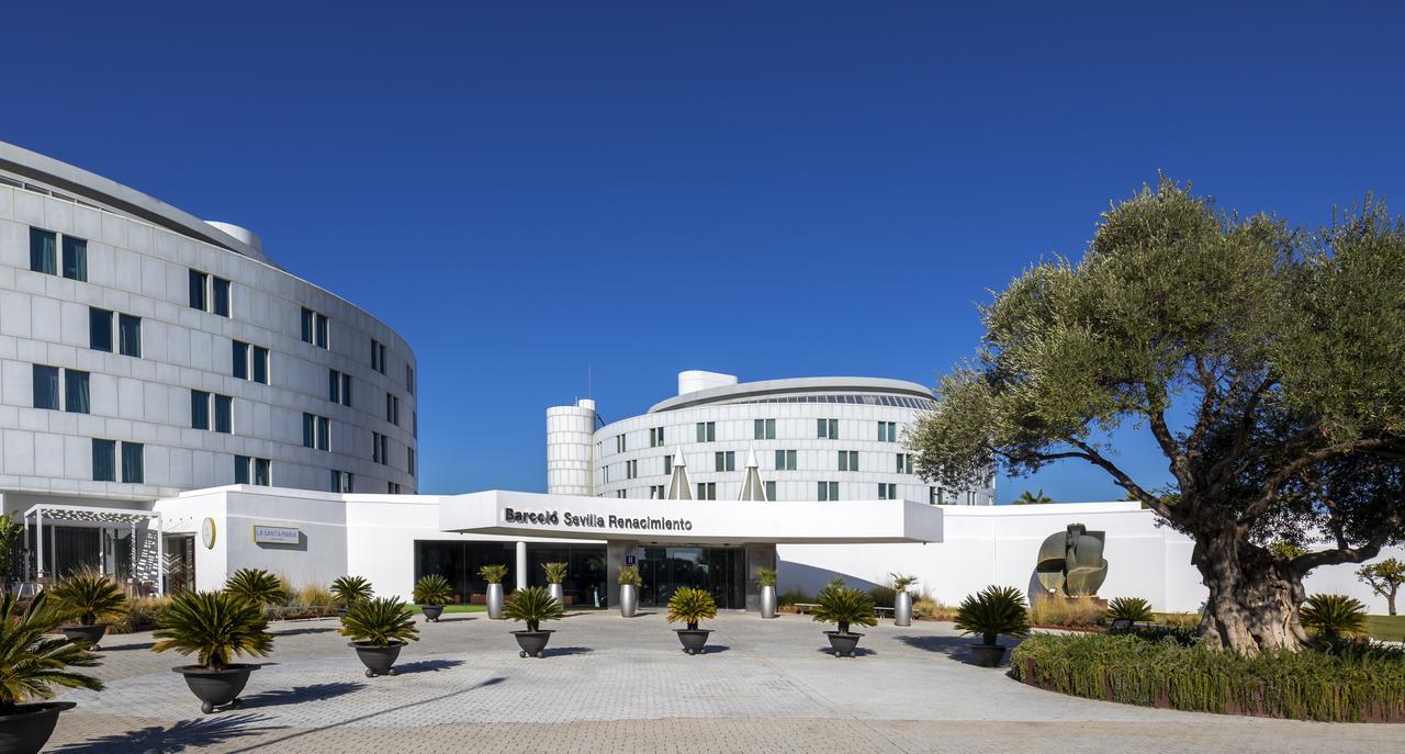 Barcelo Sevilla Renacimiento Esterno foto The photo shows a modern building complex with a sleek architectural design. There are two prominent structures with rounded shapes featuring large windows. In the foreground, there is a well-maintained entrance area with a sign that reads "Benetis S
