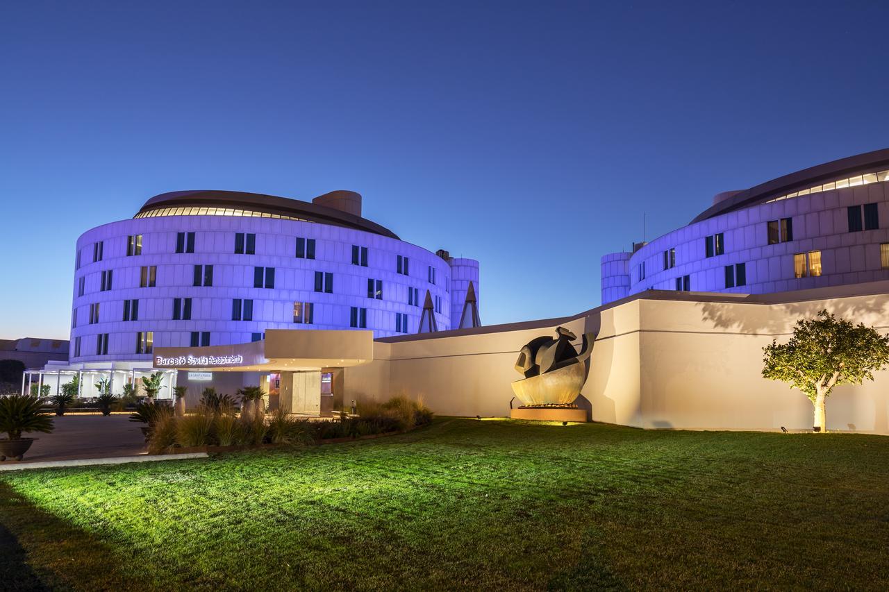 Barcelo Sevilla Renacimiento Esterno foto The photo shows a modern building complex illuminated in purple light against a twilight sky. The architectural design features curved shapes and large windows. In the foreground, there is a manicured green lawn and a sculpture prominently placed in 