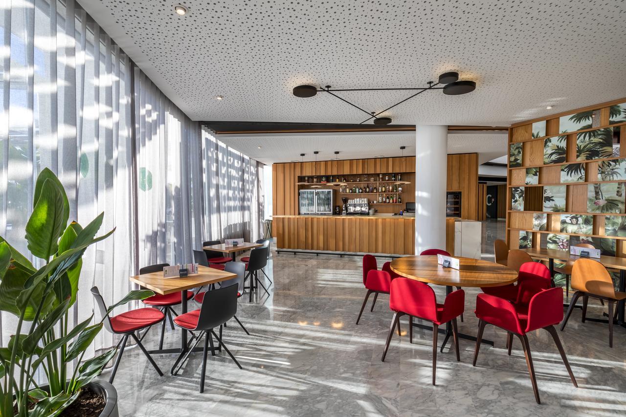 Barcelo Sevilla Renacimiento Esterno foto The photo displays a modern interior space, likely a café or dining area. There are several small round tables with red and black chairs arranged around the room. One side features large windows with sheer curtains that allow natural light to filter 