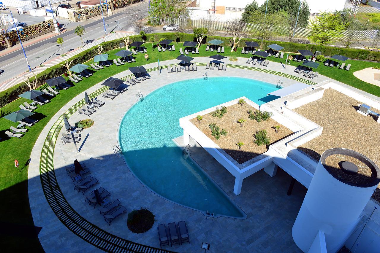 Barcelo Sevilla Renacimiento Esterno foto The photo shows a modern outdoor pool area with a circular swimming pool surrounded by lush green grass. There are several lounge chairs placed around the pool, providing a relaxing space for sunbathing. In the center of the pool area, there is a sma