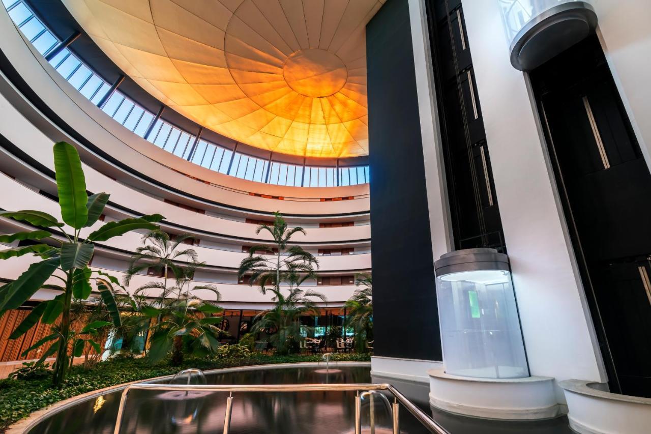 Barcelo Sevilla Renacimiento Esterno foto The photo depicts a modern architectural interior with a circular design. It features multiple levels, each surrounded by large windows that allow natural light to illuminate the space. The ceiling has a unique, dome-like structure with a warm orange