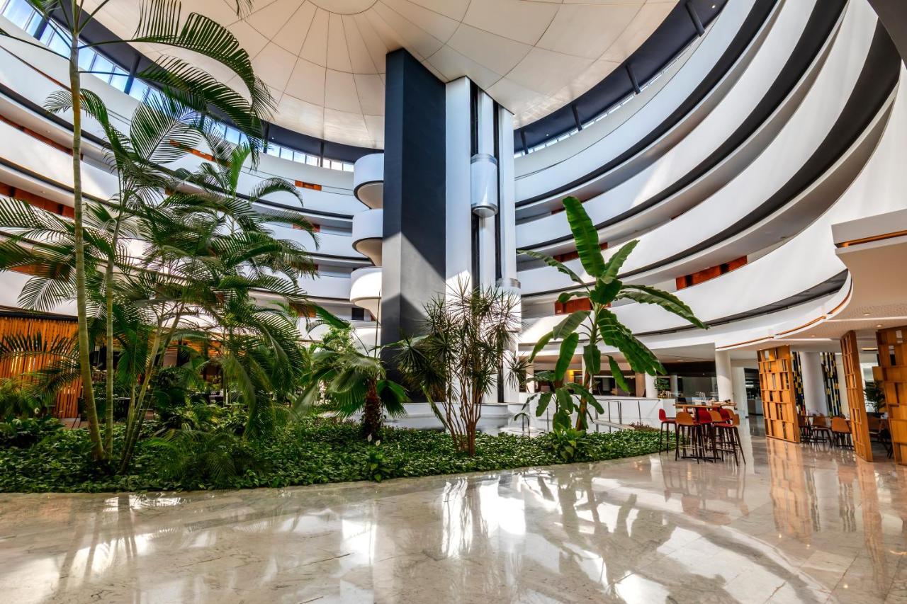 Barcelo Sevilla Renacimiento Esterno foto The photo depicts a modern atrium with a spacious, curved design. The architecture features multiple levels with smooth lines and a high ceiling. Lush greenery, including palm-like plants, is present throughout the atrium, creating a vibrant and invi