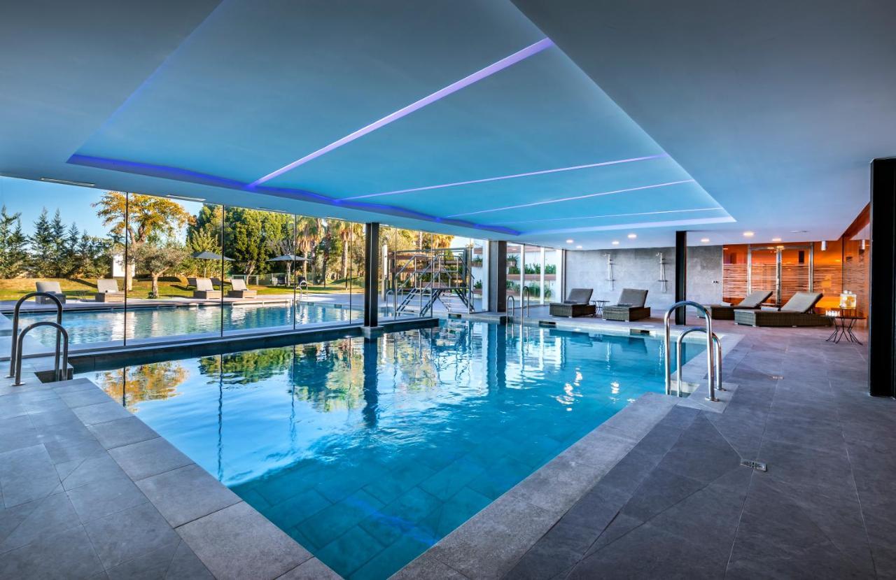 Barcelo Sevilla Renacimiento Esterno foto The photo shows a modern indoor swimming pool area. The pool has clear, blue water and is surrounded by a sleek, minimalist design. There are comfortable lounge chairs positioned nearby, and large windows provide a view of an outdoor space with green