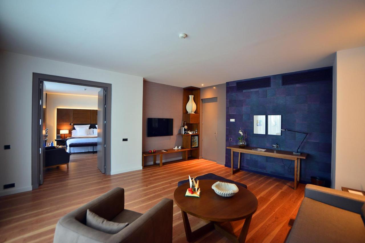 Barcelo Sevilla Renacimiento Esterno foto The photo shows a modern hotel room or suite. In the foreground, there is a seating area with two brown armchairs and a round coffee table that features decorative items. The flooring is made of light wood. On the left side, a doorway leads to anothe