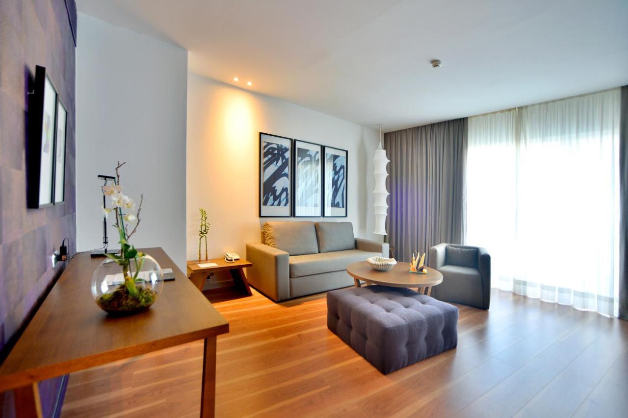 Barcelo Sevilla Renacimiento Esterno foto The photo displays a modern living room. There is a light gray sofa facing a coffee table and a plush ottoman in a similar gray hue. On the wall behind the sofa, there are three framed artworks with abstract designs. A large window with sheer curtain
